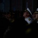 A group of military personnel are gathered in a dimly lit indoor or semi-outdoor setting, looking upward attentively. Two senior naval officers in white dress uniforms with gold insignia are prominently visible in the foreground, surrounded by other uniformed personnel in darker camouflage attire. The scene suggests a formal military ceremony or presentation taking place at night or in low-light conditions.