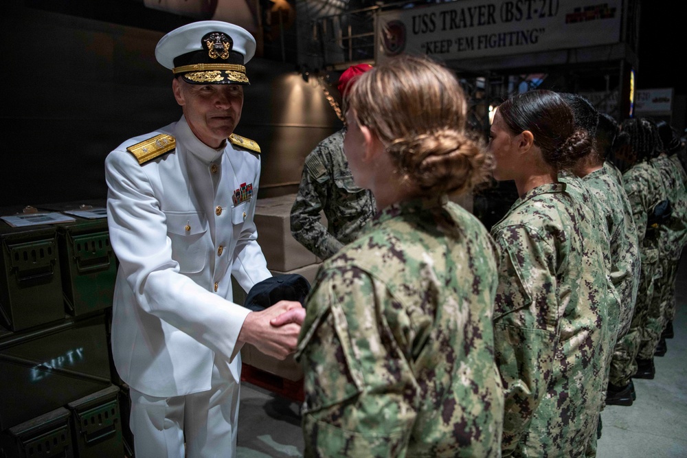A senior U.S. Navy officer in white dress uniform is shaking hands with enlisted personnel wearing Navy Working Uniform (NWU) camouflage. The setting appears to be aboard or near the USS Trayer (BST-21), a Battle Stations Training ship used in Navy boot camp at Recruit Training Command. The officer appears to be greeting or congratulating a line of recruits or sailors.