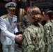 A senior U.S. Navy officer in white dress uniform is shaking hands with enlisted personnel wearing Navy Working Uniform (NWU) camouflage. The setting appears to be aboard or near the USS Trayer (BST-21), a Battle Stations Training ship used in Navy boot camp at Recruit Training Command. The officer appears to be greeting or congratulating a line of recruits or sailors.