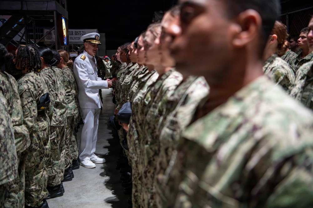 A senior naval officer in white dress uniform is addressing or inspecting rows of enlisted sailors wearing Navy Working Uniform (NWU) camouflage. The formation appears to be taking place in a large indoor or covered facility, with a sign partially visible in the background reading 'BST-21' and 'FIGHTIN'. The scene suggests a formal military ceremony, inspection, or graduation event.