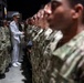 A senior naval officer in white dress uniform is addressing or inspecting rows of enlisted sailors wearing Navy Working Uniform (NWU) camouflage. The formation appears to be taking place in a large indoor or covered facility, with a sign partially visible in the background reading 'BST-21' and 'FIGHTIN'. The scene suggests a formal military ceremony, inspection, or graduation event.