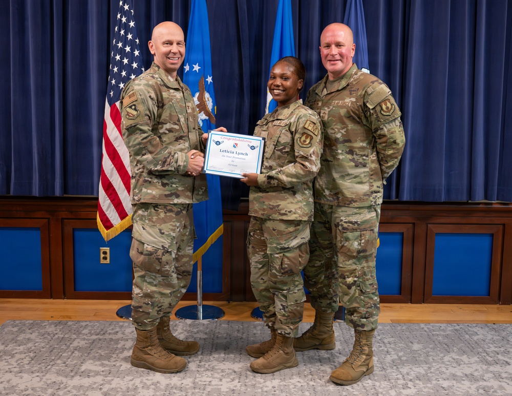 Three U.S. Air Force personnel in OCP (Operational Camouflage Pattern) uniforms pose for a formal promotion or award ceremony photo. The central figure, identified on the certificate as 'Leticia Lynch,' holds a congratulatory certificate while shaking hands with one of the flanking service members. The setting appears to be an official military ceremony room with U.S. and Air Force flags in the background.
