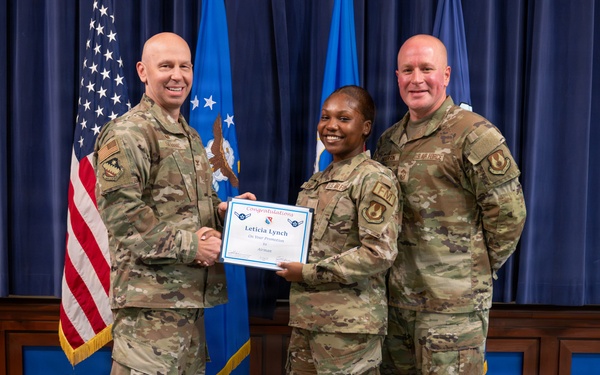 Three U.S. Air Force personnel in OCP (Operational Camouflage Pattern) uniforms pose for a formal promotion or award ceremony photo. The central figure, identified on the certificate as 'Leticia Lynch,' holds a congratulatory certificate while shaking hands with one of the flanking service members. The setting appears to be an official military ceremony room with U.S. and Air Force flags in the background.