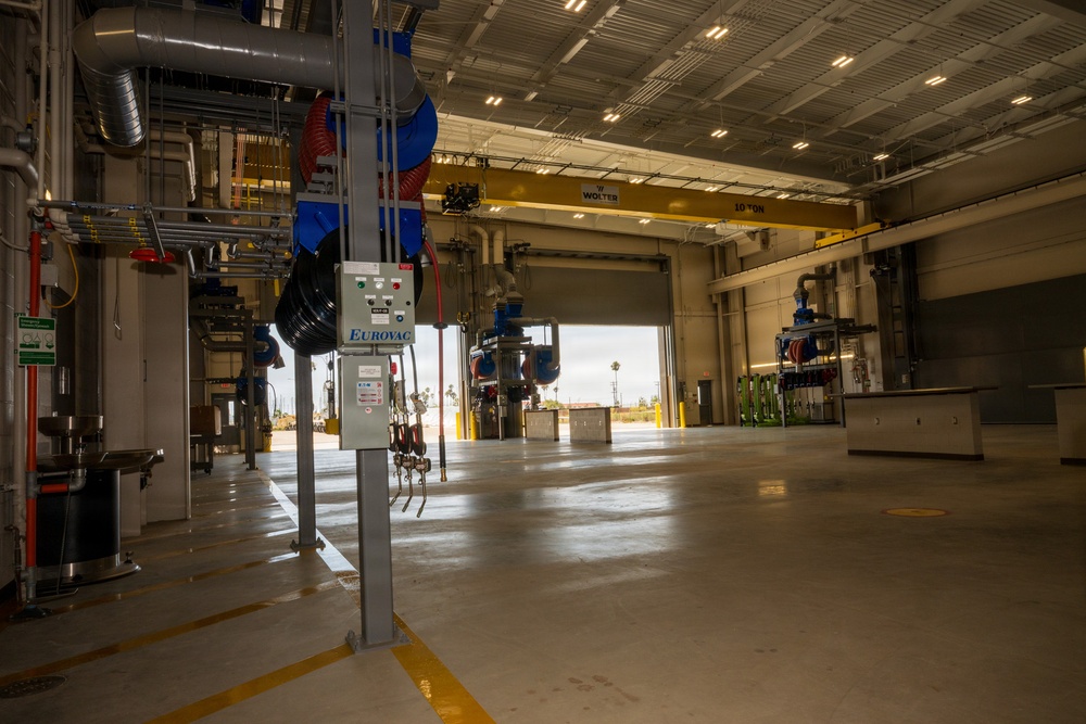 The image shows the interior of a modern industrial maintenance or service facility, likely a vehicle or heavy equipment maintenance bay. The space features overhead crane infrastructure, utility distribution systems, and specialized equipment mounted on structural columns. The facility appears newly constructed or recently commissioned, with a clean epoxy-coated concrete floor and an open bay door revealing an outdoor area.