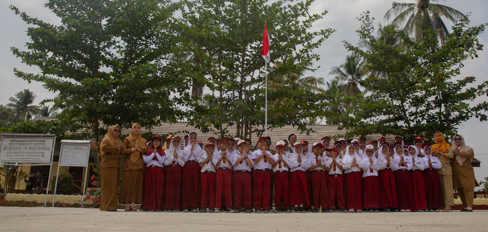 A group of Indonesian elementary school students in red-and-white uniforms are posed in a line performing a traditional greeting gesture (hands pressed together) in front of their school. Two adult teachers or staff members stand at either end of the student line. A sign board and Indonesian flag (Merah Putih) are visible in the background.