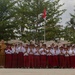 A group of Indonesian elementary school students in red-and-white uniforms are posed in a line performing a traditional greeting gesture (hands pressed together) in front of their school. Two adult teachers or staff members stand at either end of the student line. A sign board and Indonesian flag (Merah Putih) are visible in the background.