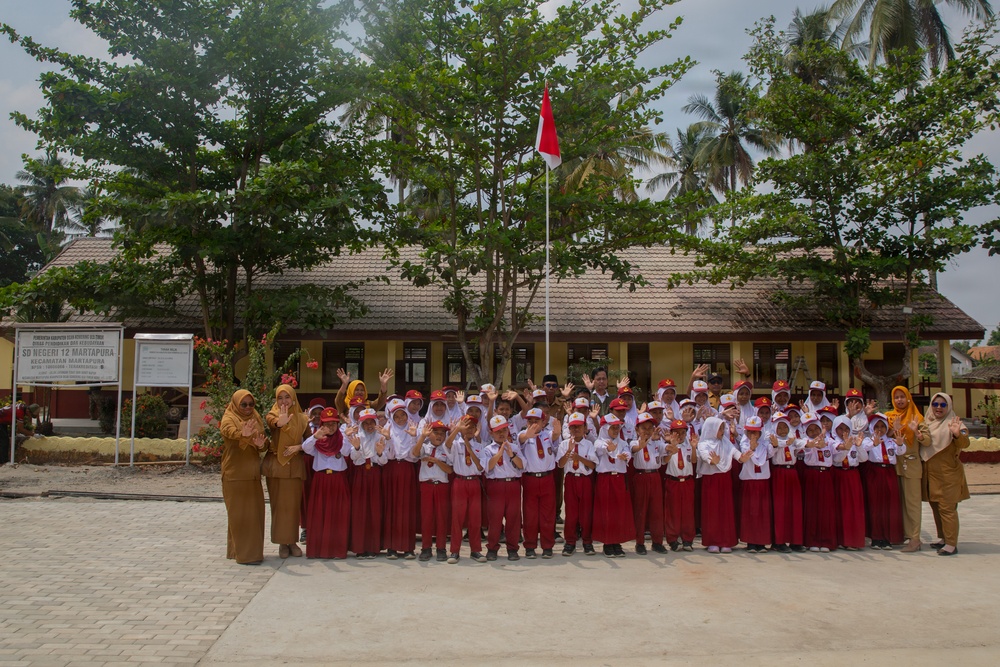 A group photo taken at SD Negeri 12 Martapura, an Indonesian public elementary school. Students dressed in the standard Indonesian school uniform of white tops and red bottoms are posing with teachers in front of the school building. The children and adults are waving at the camera in a celebratory manner.