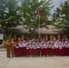 A group photo taken at SD Negeri 12 Martapura, an Indonesian public elementary school. Students dressed in the standard Indonesian school uniform of white tops and red bottoms are posing with teachers in front of the school building. The children and adults are waving at the camera in a celebratory manner.