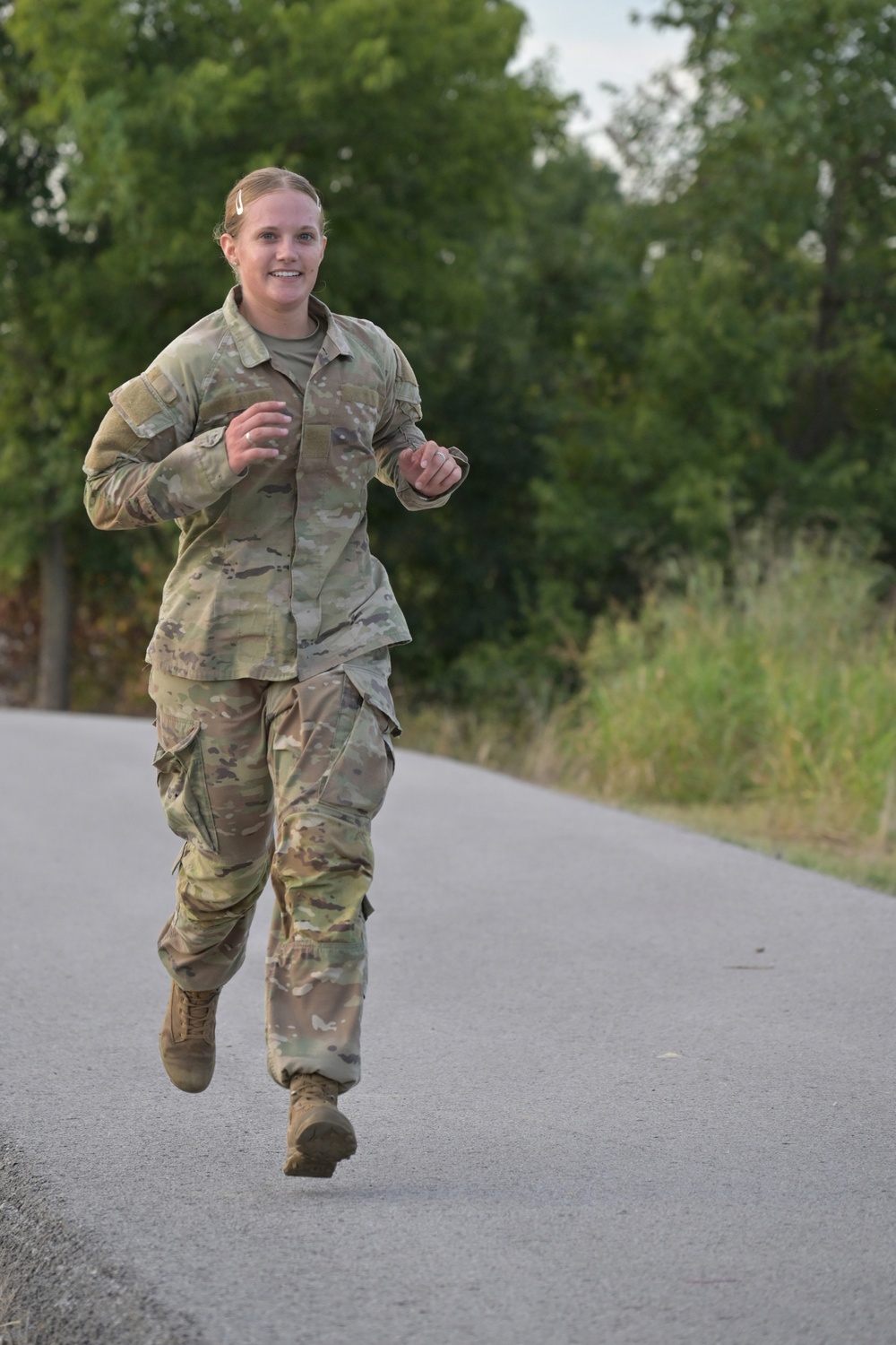 A female military service member in OCP (Operational Camouflage Pattern) uniform is running along a paved road. She appears to be smiling and in good physical condition, suggesting a physical training or fitness test event. The background features lush green trees and vegetation consistent with a warm-season outdoor setting.
