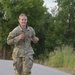A female military service member in OCP (Operational Camouflage Pattern) uniform is running along a paved road. She appears to be smiling and in good physical condition, suggesting a physical training or fitness test event. The background features lush green trees and vegetation consistent with a warm-season outdoor setting.