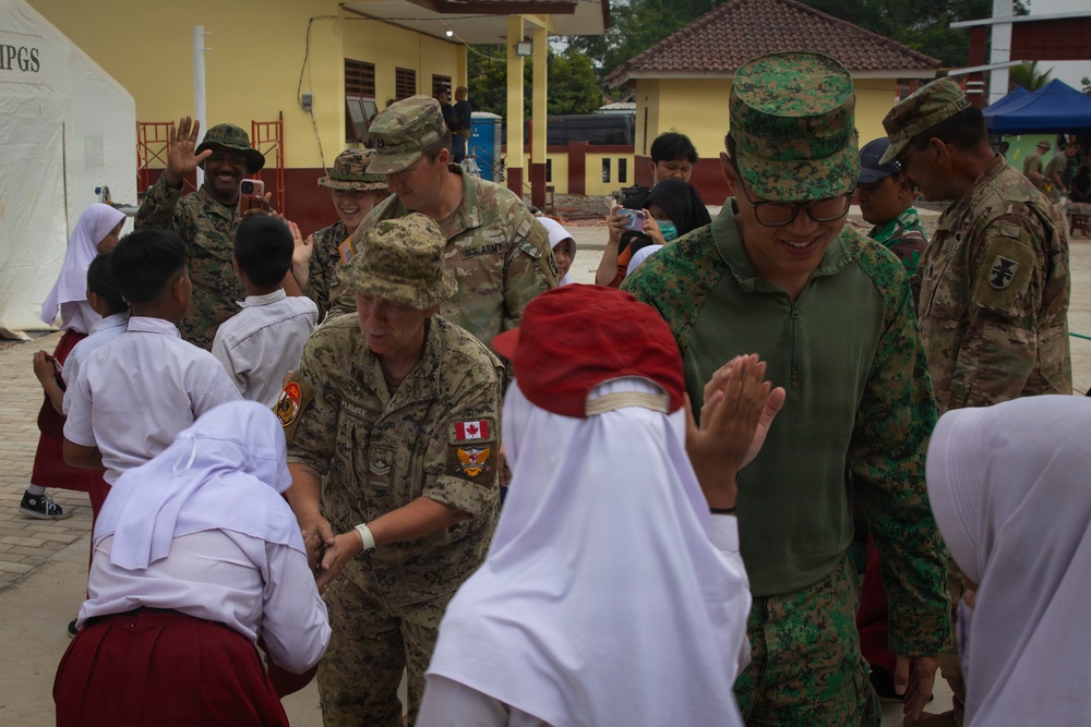 Military personnel from multiple nations are interacting with local schoolchildren in what appears to be a community engagement or humanitarian assistance event. The soldiers, wearing uniforms from at least Canada, the United States, and Singapore, are greeting and playing with children dressed in school uniforms including white hijabs and red skirts. The setting appears to be a school or community compound in Southeast Asia, likely Indonesia.