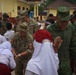 Military personnel from multiple nations are interacting with local schoolchildren in what appears to be a community engagement or humanitarian assistance event. The soldiers, wearing uniforms from at least Canada, the United States, and Singapore, are greeting and playing with children dressed in school uniforms including white hijabs and red skirts. The setting appears to be a school or community compound in Southeast Asia, likely Indonesia.