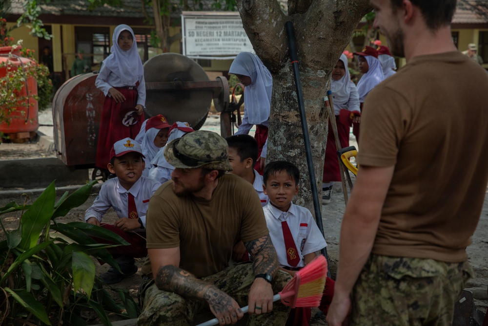 The image shows military personnel in olive/tan uniforms interacting with Indonesian schoolchildren at what appears to be SD Negeri 12 Martapura, a primary school in Indonesia. The children are wearing standard Indonesian school uniforms consisting of white shirts, red shorts/skirts, and red ties with white caps. The scene appears to depict a humanitarian assistance or community engagement activity.