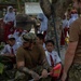 The image shows military personnel in olive/tan uniforms interacting with Indonesian schoolchildren at what appears to be SD Negeri 12 Martapura, a primary school in Indonesia. The children are wearing standard Indonesian school uniforms consisting of white shirts, red shorts/skirts, and red ties with white caps. The scene appears to depict a humanitarian assistance or community engagement activity.