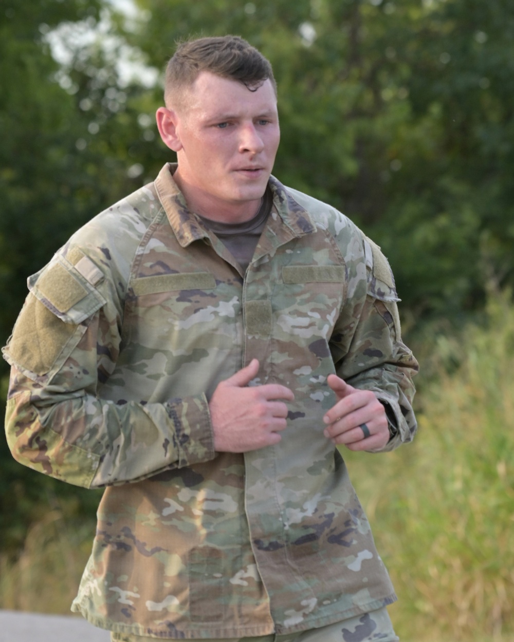 A male soldier in US Army OCP (Operational Camouflage Pattern) uniform is captured mid-stride, appearing to be running or jogging. He is wearing a black ring on his right hand and has a subdued flag patch on his left shoulder. The background consists of green trees and vegetation, suggesting an outdoor training or fitness environment.