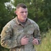 A male soldier in US Army OCP (Operational Camouflage Pattern) uniform is captured mid-stride, appearing to be running or jogging. He is wearing a black ring on his right hand and has a subdued flag patch on his left shoulder. The background consists of green trees and vegetation, suggesting an outdoor training or fitness environment.