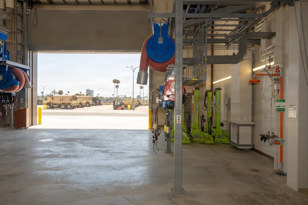 The image shows the interior of a modern vehicle maintenance facility or motor pool garage with an open bay door revealing a paved yard with military-style trucks and heavy equipment parked outside. The facility is well-equipped with retractable hose reels, green hydraulic vehicle lifts, overhead piping, and utility connections mounted on structural columns. The setting appears to be a military or government installation based on the tan/desert-colored tactical vehicles visible in the background.