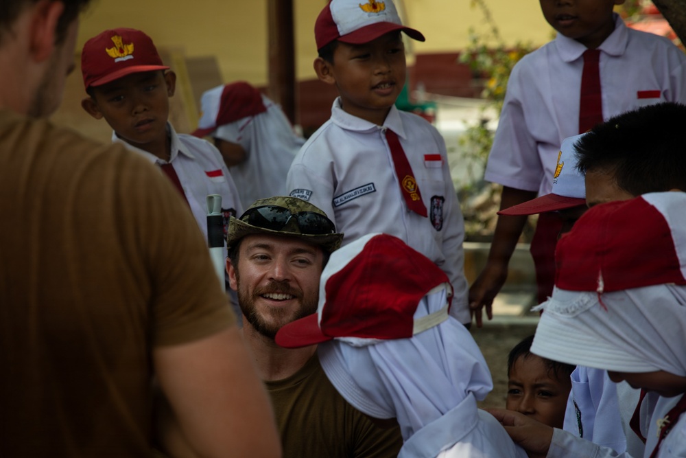 A smiling Western adult male wearing a camouflage cap and olive/tan shirt is crouching among a group of young school children. The children are wearing matching white uniforms with red ties and red-and-white caps, consistent with Indonesian elementary school attire. The setting appears to be an outdoor school courtyard, suggesting a community visit or humanitarian engagement.