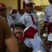 A smiling Western adult male wearing a camouflage cap and olive/tan shirt is crouching among a group of young school children. The children are wearing matching white uniforms with red ties and red-and-white caps, consistent with Indonesian elementary school attire. The setting appears to be an outdoor school courtyard, suggesting a community visit or humanitarian engagement.