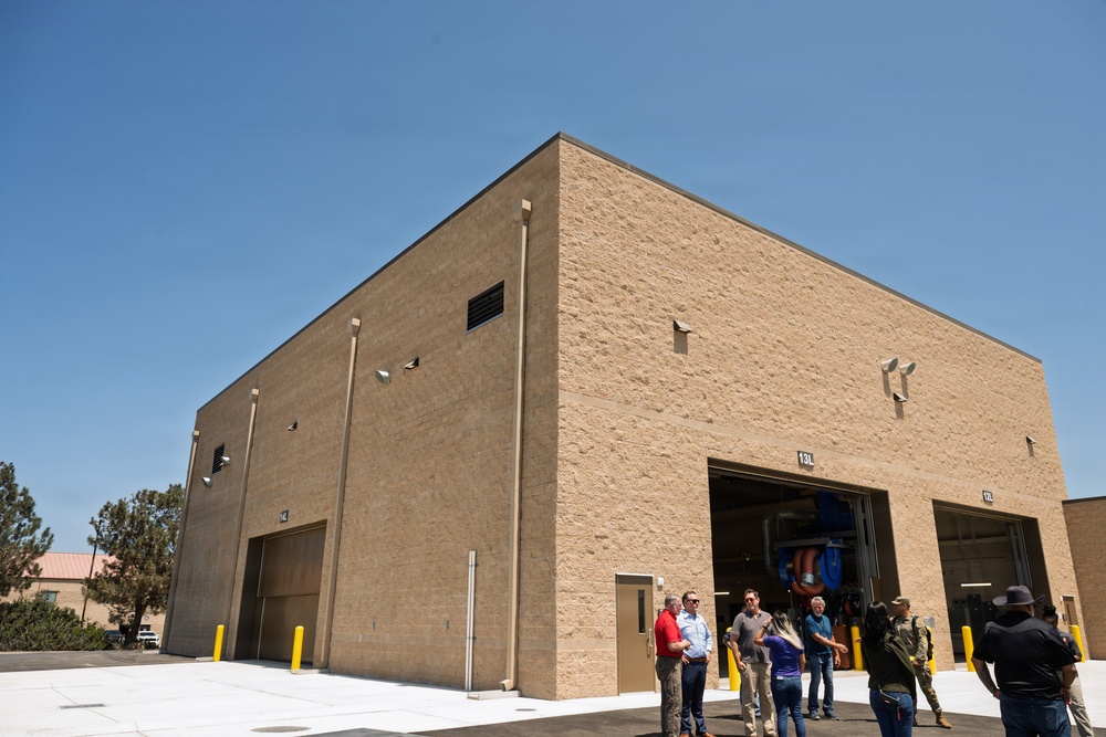 A large, newly constructed tan/beige masonry building with multiple large bay doors, consistent with a maintenance facility, fire station, or military/government garage. A group of approximately 8-10 people are gathered near the entrance, appearing to be on a tour or inspection of the facility. The building appears modern and well-maintained, set in a sunny, arid environment.