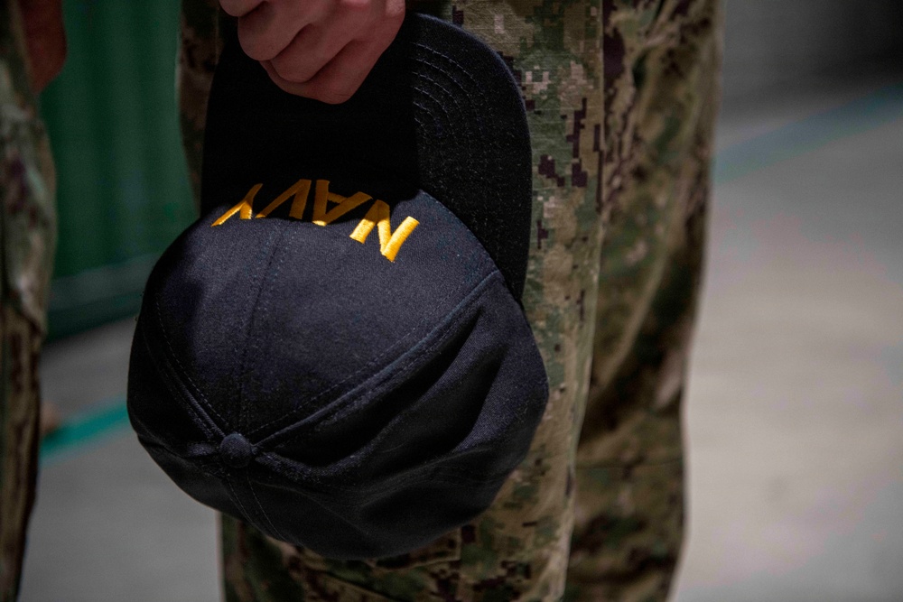 A close-up photograph showing a person holding a black Navy cap with gold embroidered lettering reading 'NAVY'. The individual is wearing digital camouflage uniform trousers consistent with U.S. Navy Working Uniform (NWU) Type III pattern. The cap is held at the side, facing outward toward the viewer.