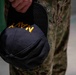 A close-up photograph showing a person holding a black Navy cap with gold embroidered lettering reading 'NAVY'. The individual is wearing digital camouflage uniform trousers consistent with U.S. Navy Working Uniform (NWU) Type III pattern. The cap is held at the side, facing outward toward the viewer.