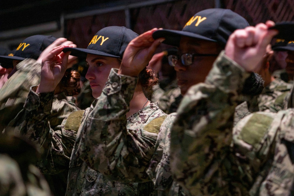 A group of U.S. Navy recruits or personnel are standing in formation while simultaneously placing or adjusting dark navy blue caps with gold 'NAVY' lettering on their heads. They are wearing U.S. Navy Working Uniform (NWU) Type III camouflage pattern uniforms. The scene appears to be part of a formal military ceremony or training exercise.