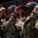 A group of U.S. Navy recruits or personnel are standing in formation while simultaneously placing or adjusting dark navy blue caps with gold 'NAVY' lettering on their heads. They are wearing U.S. Navy Working Uniform (NWU) Type III camouflage pattern uniforms. The scene appears to be part of a formal military ceremony or training exercise.