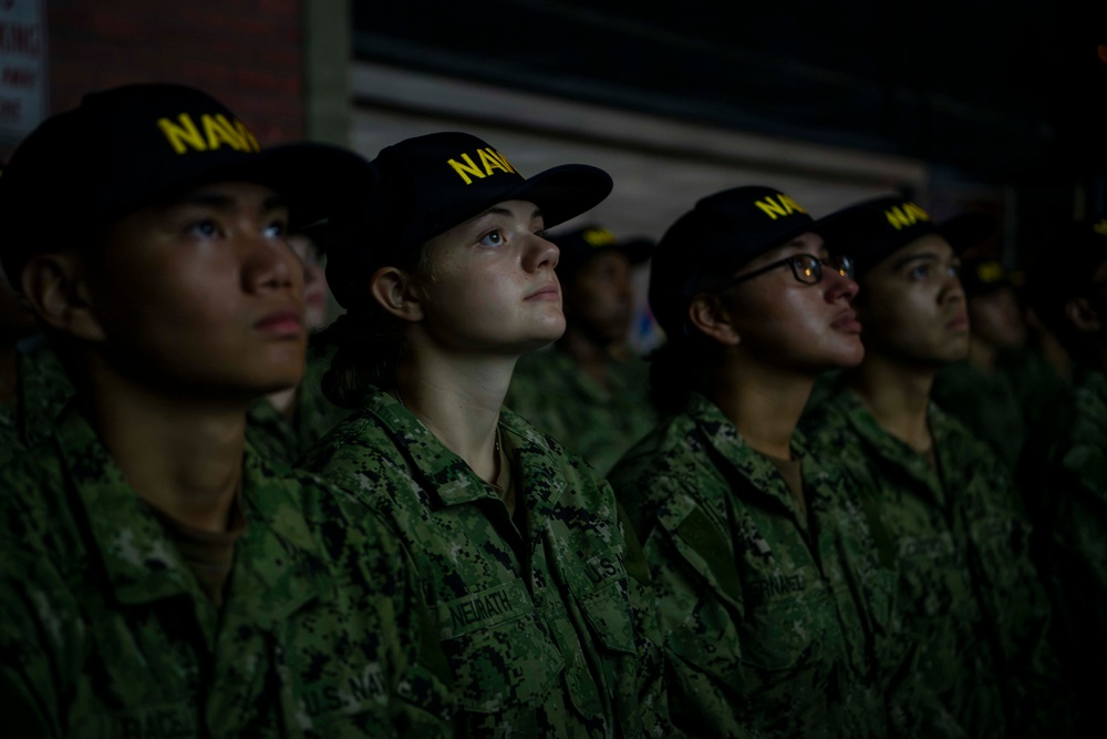 A group of U.S. Navy personnel standing in formation, wearing matching Navy Working Uniforms (NWU Type III) and black 'NAVY' caps with yellow lettering. The subjects appear to be recruits or junior enlisted members, all looking upward in the same direction. The image is dramatically lit, with one central figure notably illuminated against a dark background.