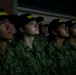 A group of U.S. Navy personnel standing in formation, wearing matching Navy Working Uniforms (NWU Type III) and black 'NAVY' caps with yellow lettering. The subjects appear to be recruits or junior enlisted members, all looking upward in the same direction. The image is dramatically lit, with one central figure notably illuminated against a dark background.