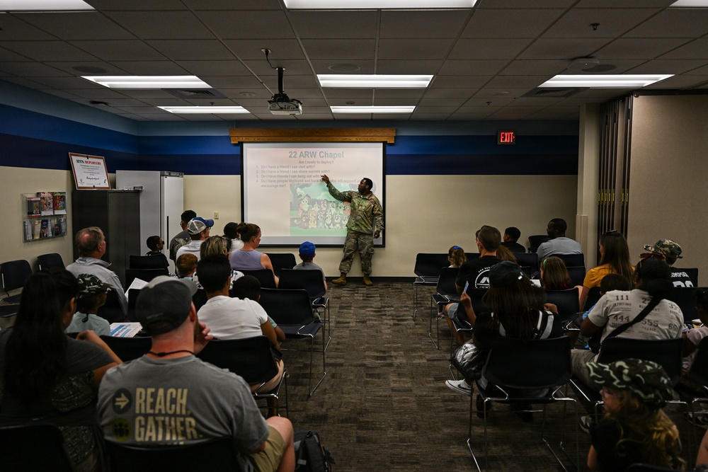 A military chaplain or officer in camouflage uniform is presenting to a mixed audience of adults and children in a conference or briefing room. The presentation slide visible on the screen reads '22 ARW Chapel' with the subtitle 'Am I ready to deploy?' The audience is seated in rows of chairs facing the presenter and projection screen.