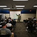A military chaplain or officer in camouflage uniform is presenting to a mixed audience of adults and children in a conference or briefing room. The presentation slide visible on the screen reads '22 ARW Chapel' with the subtitle 'Am I ready to deploy?' The audience is seated in rows of chairs facing the presenter and projection screen.
