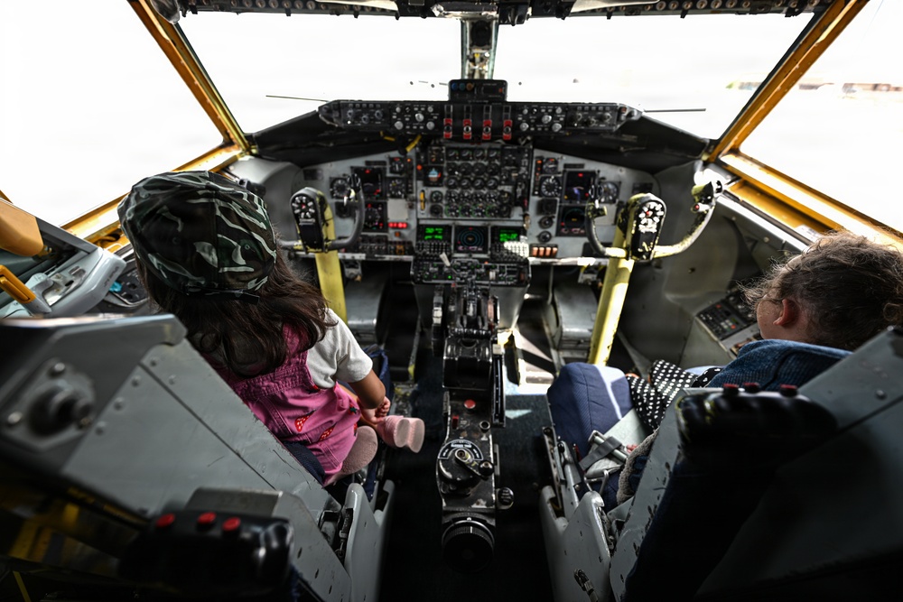 Two individuals — a young child and an adult — are seated in the cockpit of what appears to be a vintage or classic aircraft, likely on static display or during a public tour event. The child, wearing a camouflage hat and pink overalls, sits in the left pilot seat, while the adult occupies the right seat. The cockpit features an extensive analog instrument panel with numerous gauges, switches, and controls consistent with mid-20th century aviation technology.