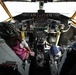 Two individuals — a young child and an adult — are seated in the cockpit of what appears to be a vintage or classic aircraft, likely on static display or during a public tour event. The child, wearing a camouflage hat and pink overalls, sits in the left pilot seat, while the adult occupies the right seat. The cockpit features an extensive analog instrument panel with numerous gauges, switches, and controls consistent with mid-20th century aviation technology.