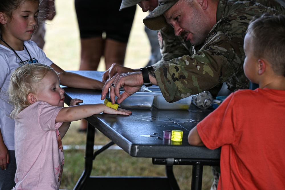 A military service member in MultiCam/OCP camouflage uniform is engaged in a hands-on activity with young children at an outdoor event table. He appears to be applying something with a sponge to a small child's outstretched hand, likely face or hand painting. The setting suggests a family-friendly military community event or open house.