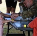 A military service member in MultiCam/OCP camouflage uniform is engaged in a hands-on activity with young children at an outdoor event table. He appears to be applying something with a sponge to a small child's outstretched hand, likely face or hand painting. The setting suggests a family-friendly military community event or open house.