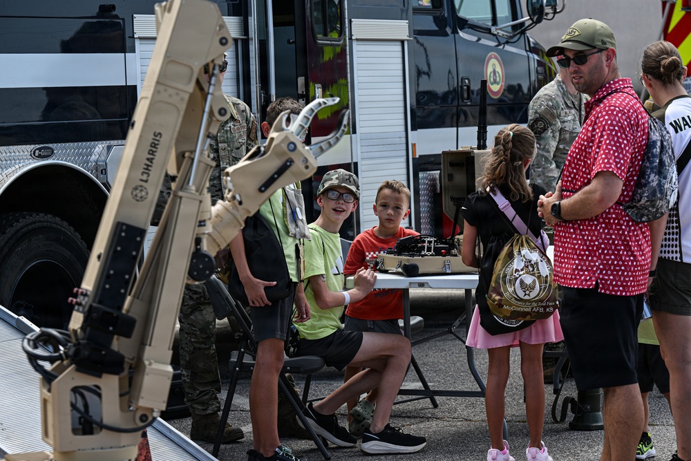 A public military open house or air show event, likely at McConnell AFB in Kansas, where children and adults are interacting with military equipment displays. A robotic arm system branded 'L3HARRIS' is prominently featured in the foreground, with children seated at a table operating or examining equipment. Military personnel in camouflage uniforms are present alongside civilians.