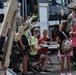 A public military open house or air show event, likely at McConnell AFB in Kansas, where children and adults are interacting with military equipment displays. A robotic arm system branded 'L3HARRIS' is prominently featured in the foreground, with children seated at a table operating or examining equipment. Military personnel in camouflage uniforms are present alongside civilians.