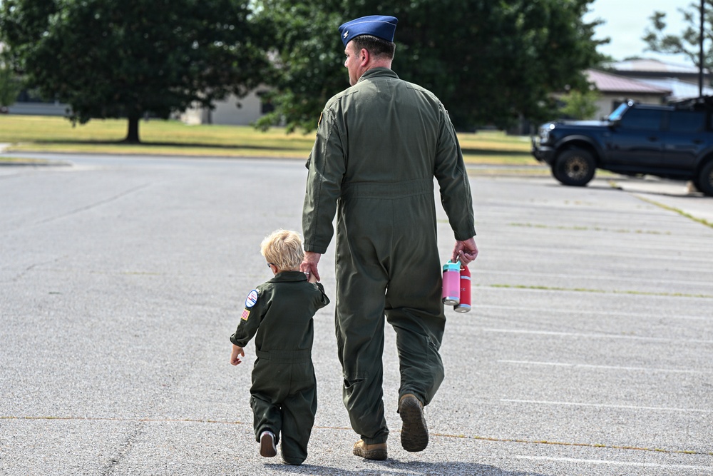 An adult military personnel member in an olive green flight suit and blue service cap walks hand-in-hand with a young child dressed in a matching miniature flight suit. Both are walking away from the camera across a paved parking area on what appears to be a military base. The adult is carrying a pink and red water bottle in their free hand.