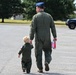 An adult military personnel member in an olive green flight suit and blue service cap walks hand-in-hand with a young child dressed in a matching miniature flight suit. Both are walking away from the camera across a paved parking area on what appears to be a military base. The adult is carrying a pink and red water bottle in their free hand.