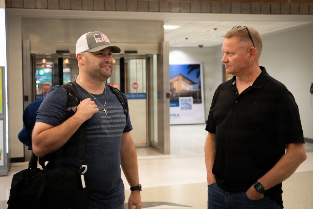Two adult males are engaged in conversation in what appears to be an airport terminal or similar transit facility. The man on the left is carrying luggage and wearing a white trucker cap with an American flag patch, while the man on the right is dressed in a black polo shirt with sunglasses resting on his head. The setting suggests an arrival or departure scenario.