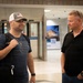 Two adult males are engaged in conversation in what appears to be an airport terminal or similar transit facility. The man on the left is carrying luggage and wearing a white trucker cap with an American flag patch, while the man on the right is dressed in a black polo shirt with sunglasses resting on his head. The setting suggests an arrival or departure scenario.
