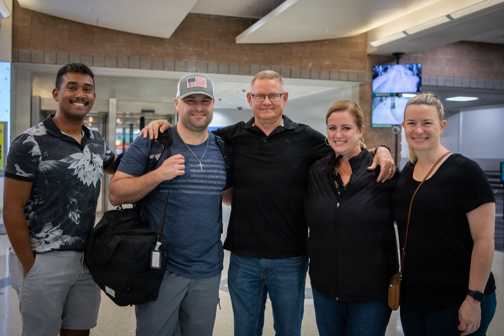 Five adults are posing together for a group photo inside what appears to be an airport terminal. They are smiling and standing close together, with some placing arms around each other's shoulders. The setting suggests a greeting or farewell occasion at the airport.