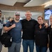 Five adults are posing together for a group photo inside what appears to be an airport terminal. They are smiling and standing close together, with some placing arms around each other's shoulders. The setting suggests a greeting or farewell occasion at the airport.
