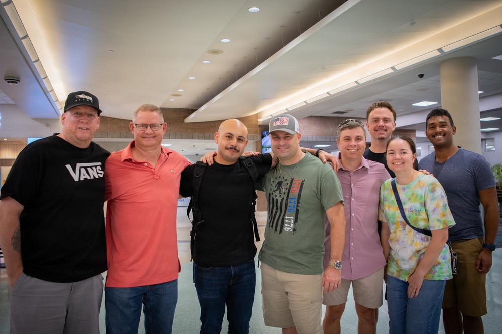 A group of eight adults poses together for a photo in what appears to be a large indoor public space, possibly an airport terminal or convention center. The group consists of seven men and one woman, smiling and standing close together in a friendly manner. The setting features modern architecture with recessed lighting and large columns.