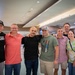 A group of eight adults poses together for a photo in what appears to be a large indoor public space, possibly an airport terminal or convention center. The group consists of seven men and one woman, smiling and standing close together in a friendly manner. The setting features modern architecture with recessed lighting and large columns.