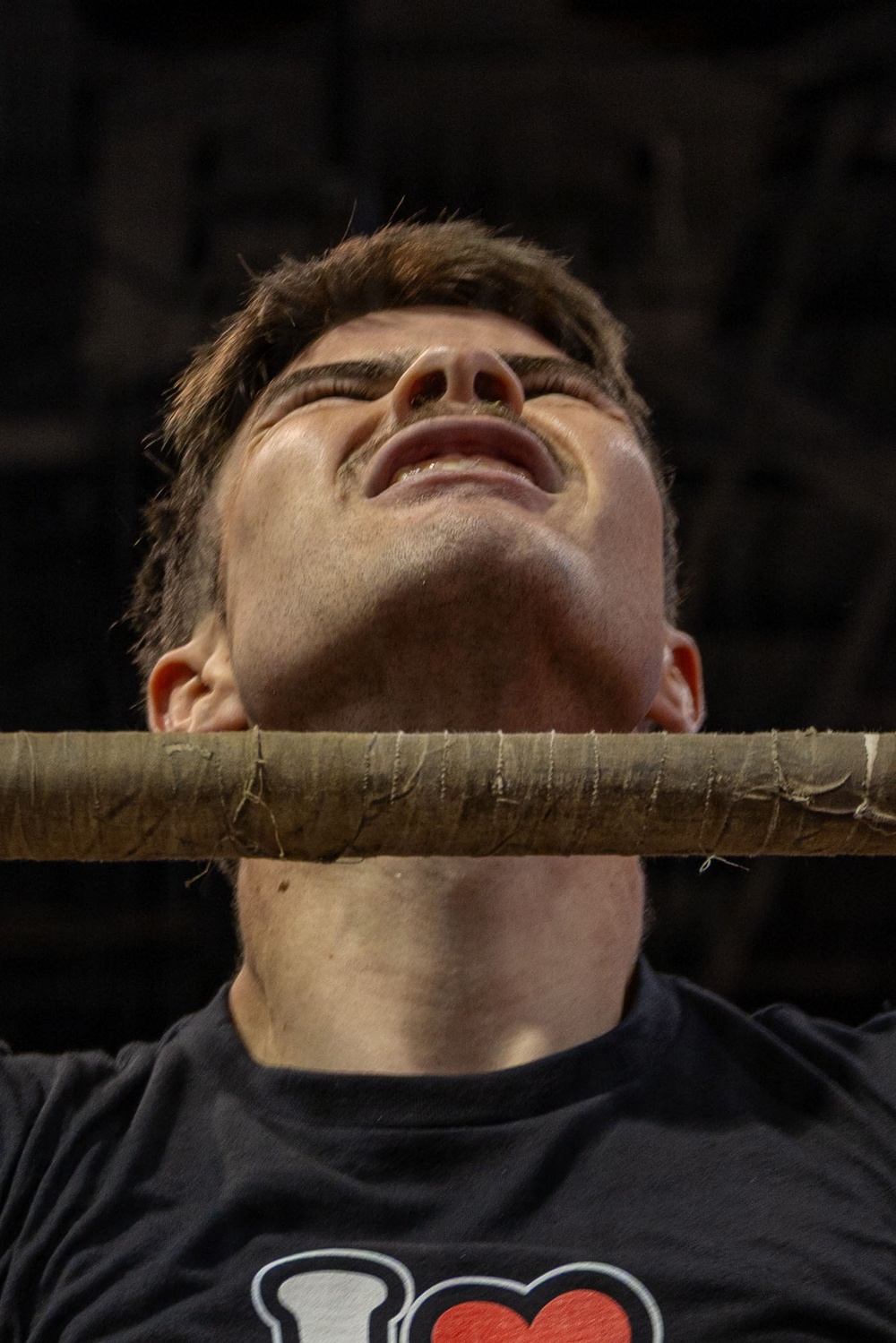 A young man is photographed from a low angle with a rope or wrapped bar pressed against his throat/neck area. His head is tilted back, eyes closed, and his expression suggests strain or discomfort. He is wearing a dark t-shirt with a partial graphic visible at the bottom.