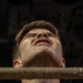 A young man is photographed from a low angle with a rope or wrapped bar pressed against his throat/neck area. His head is tilted back, eyes closed, and his expression suggests strain or discomfort. He is wearing a dark t-shirt with a partial graphic visible at the bottom.