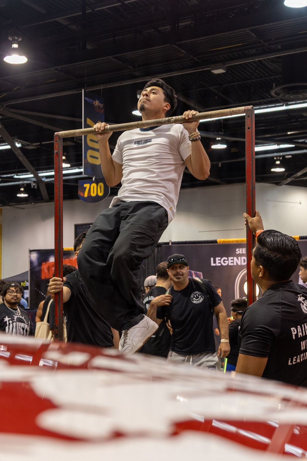 A young man is performing pull-ups on a red metal pull-up bar station at what appears to be a fitness or sports expo held in a large convention center. He is wearing a white t-shirt, dark baggy pants, and white sneakers, with a bracelet on his wrist. A crowd of spectators, many wearing black t-shirts, watches from below.