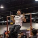 A young man is performing pull-ups on a red metal pull-up bar station at what appears to be a fitness or sports expo held in a large convention center. He is wearing a white t-shirt, dark baggy pants, and white sneakers, with a bracelet on his wrist. A crowd of spectators, many wearing black t-shirts, watches from below.